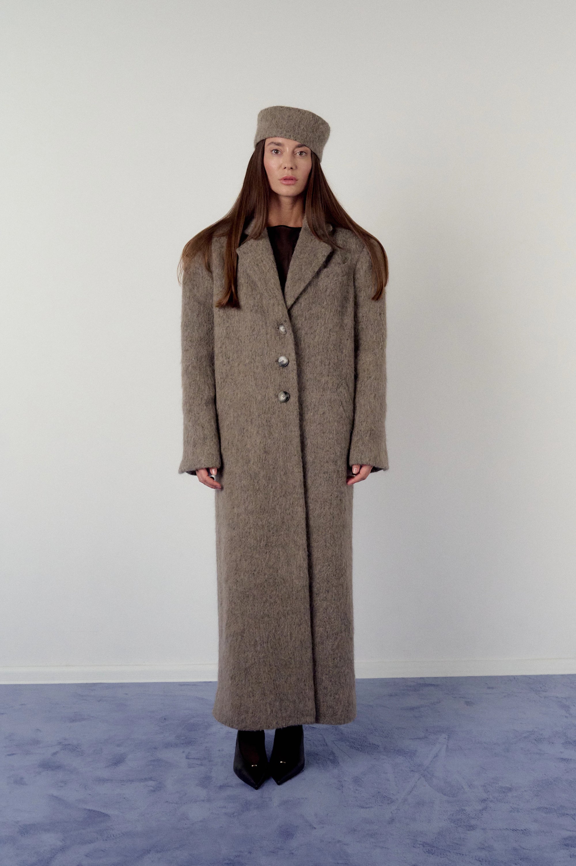 Woman wearing a long brown, warm grey coat and matching pillbox hat on a plain background