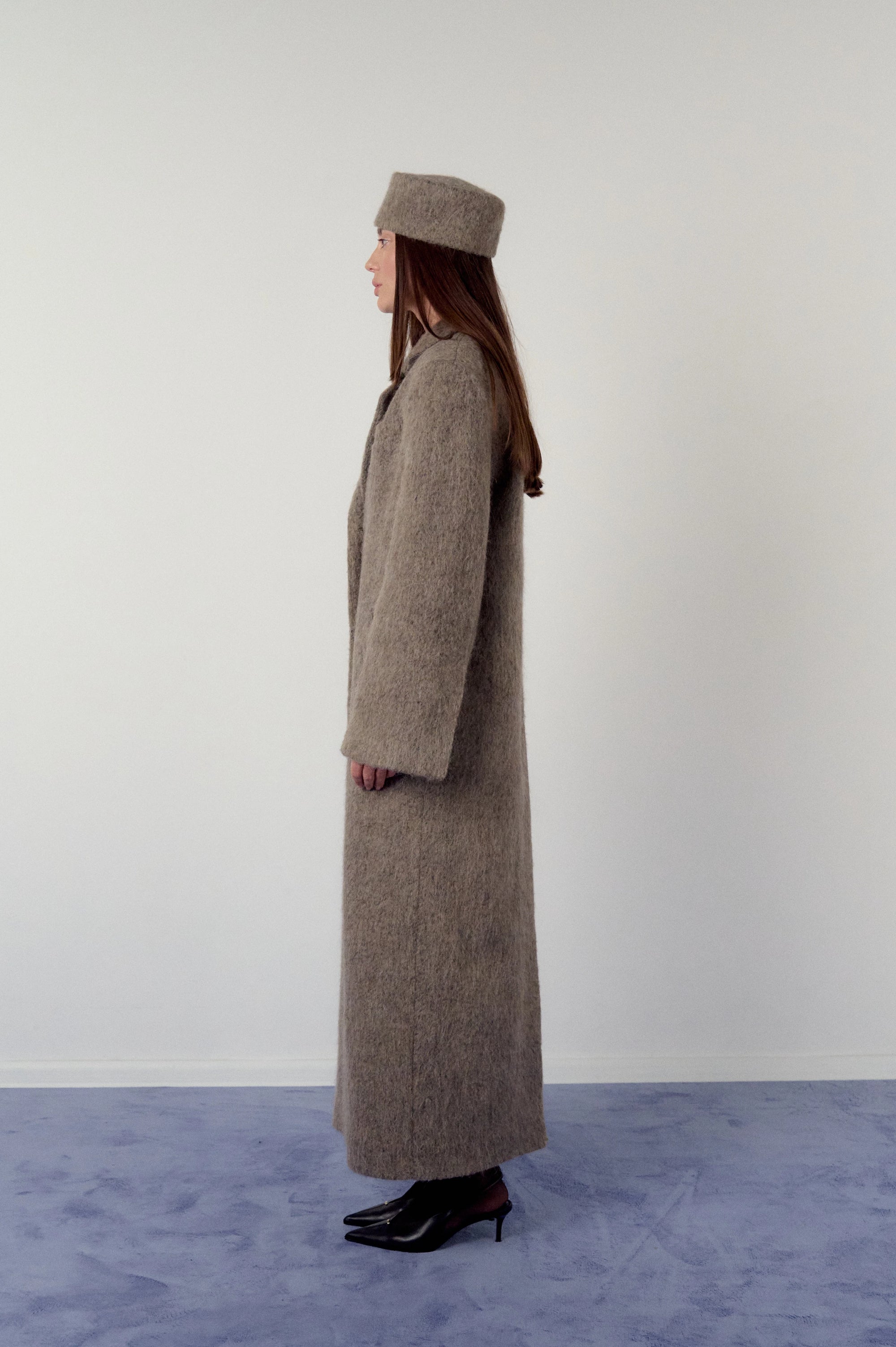 Woman wearing a long brown, warm grey coat and matching pillbox hat on a plain background