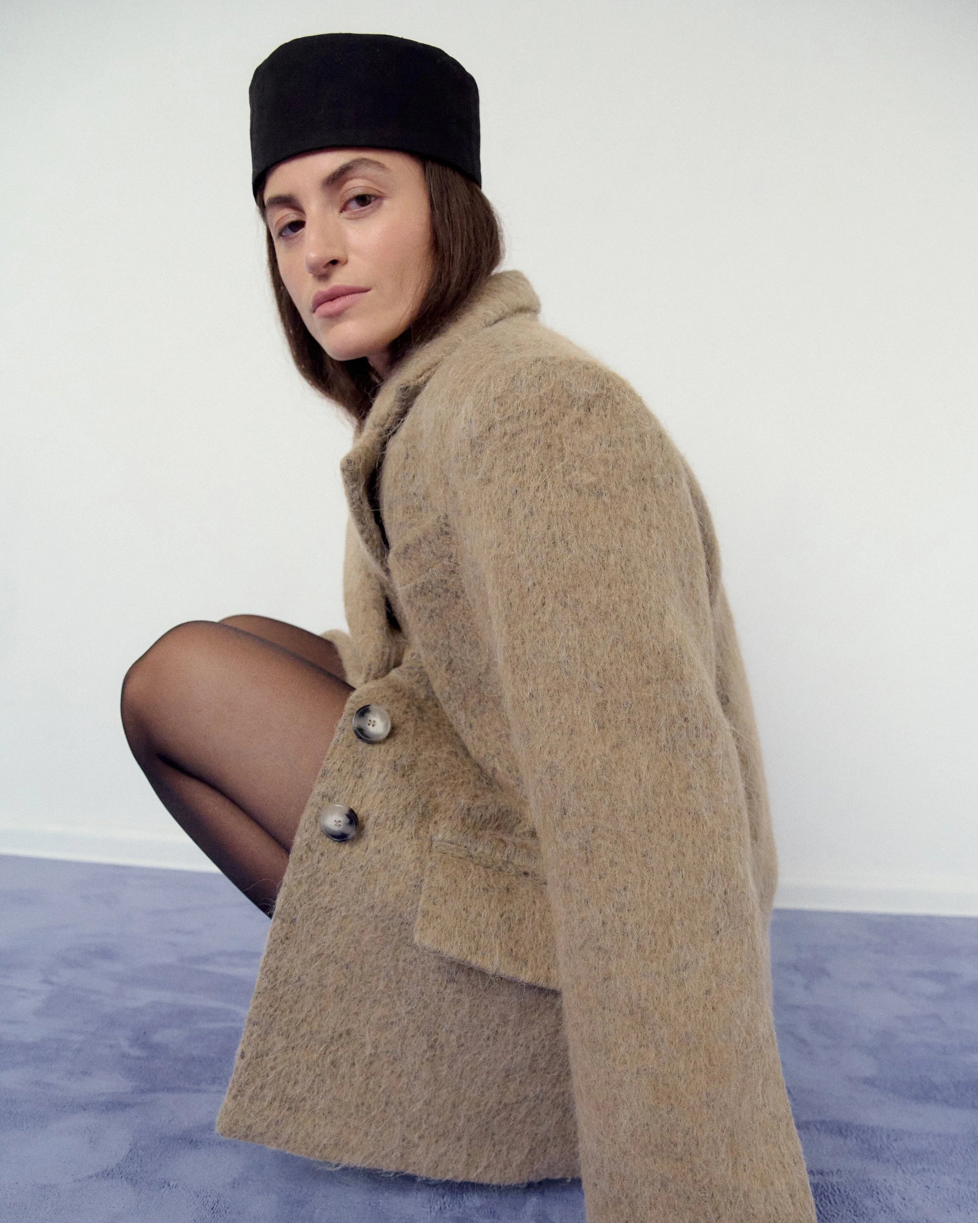 Woman wearing a beige, sand coat and black hat on a blue carpet with a white background