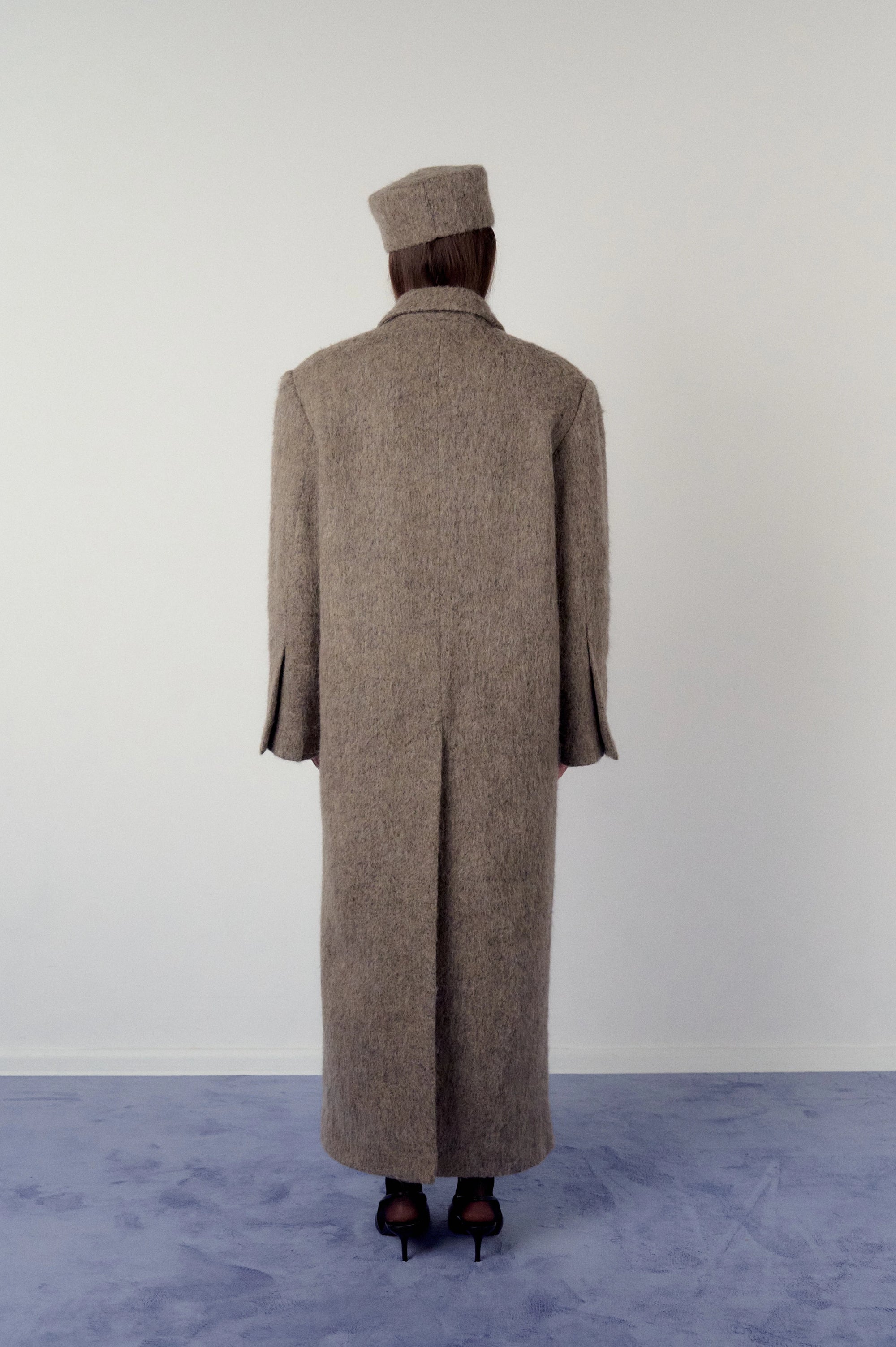 Woman wearing a long brown, warm grey coat and matching pillbox hat on a plain background