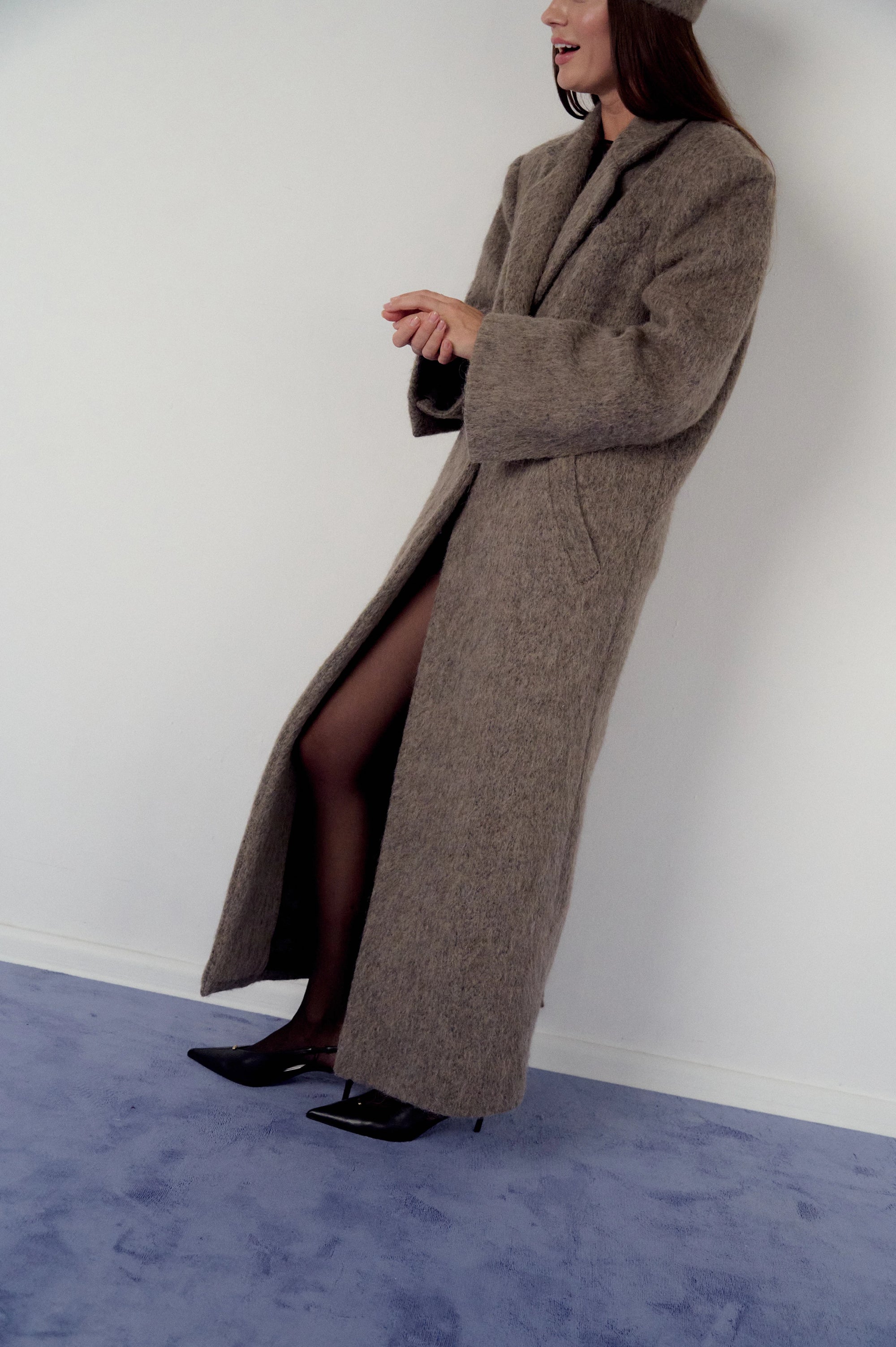 Woman wearing a long brown, warm grey coat and matching pillbox hat on a plain background