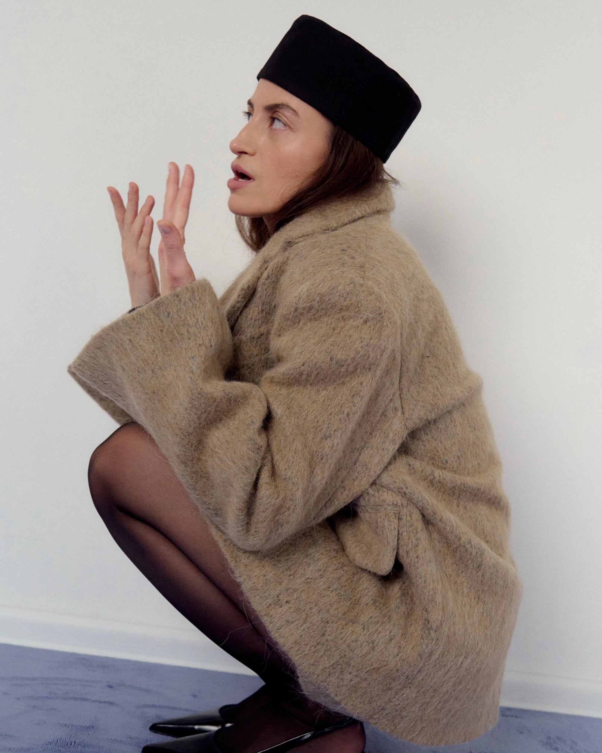 Woman in a beige, sand coat and black hat squatting on a blue carpet with a white background
