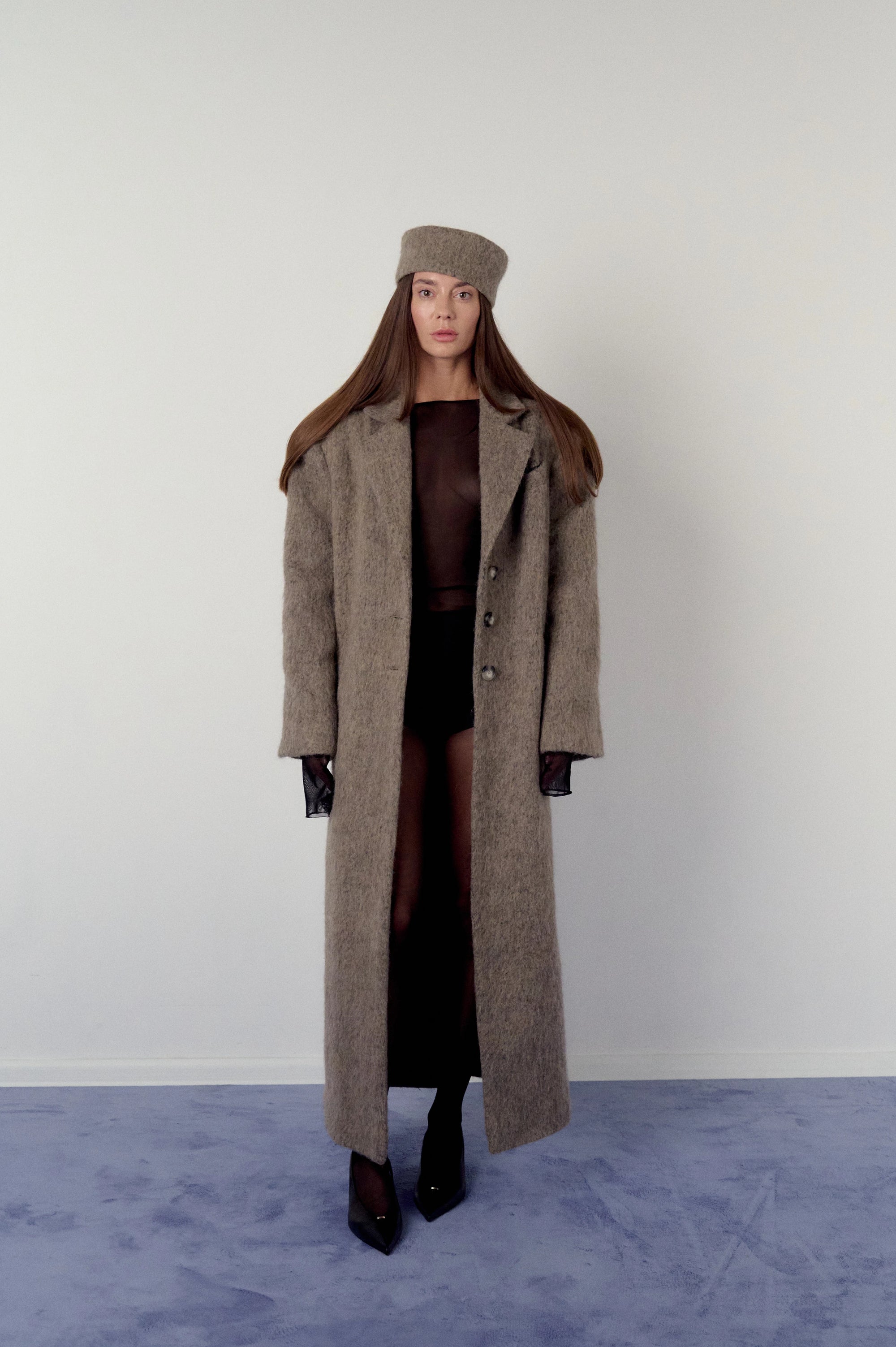 Woman wearing a long brown, warm grey coat and matching pillbox hat on a plain background