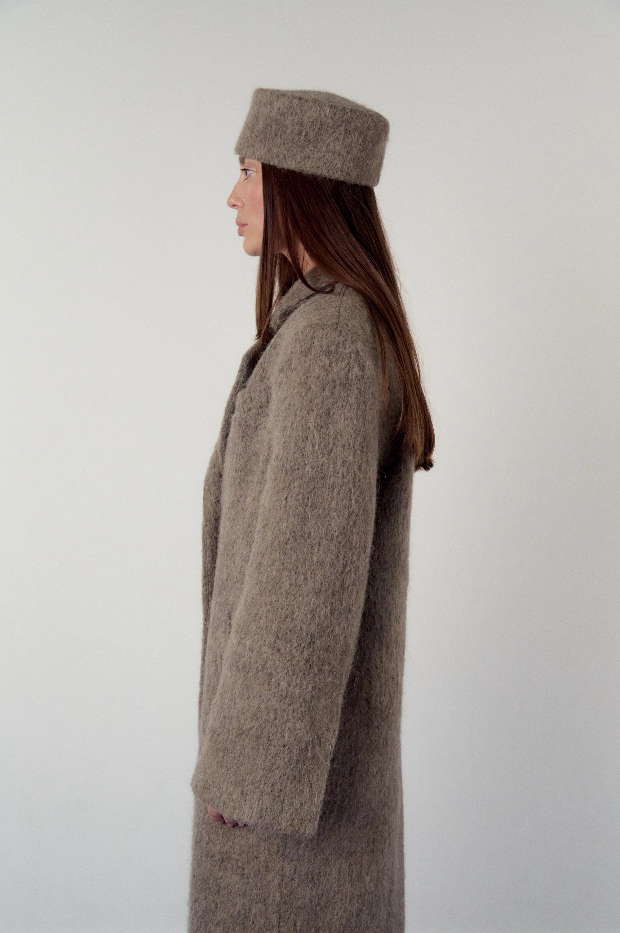 Woman wearing a long brown, warm grey coat and matching pillbox hat on a plain background