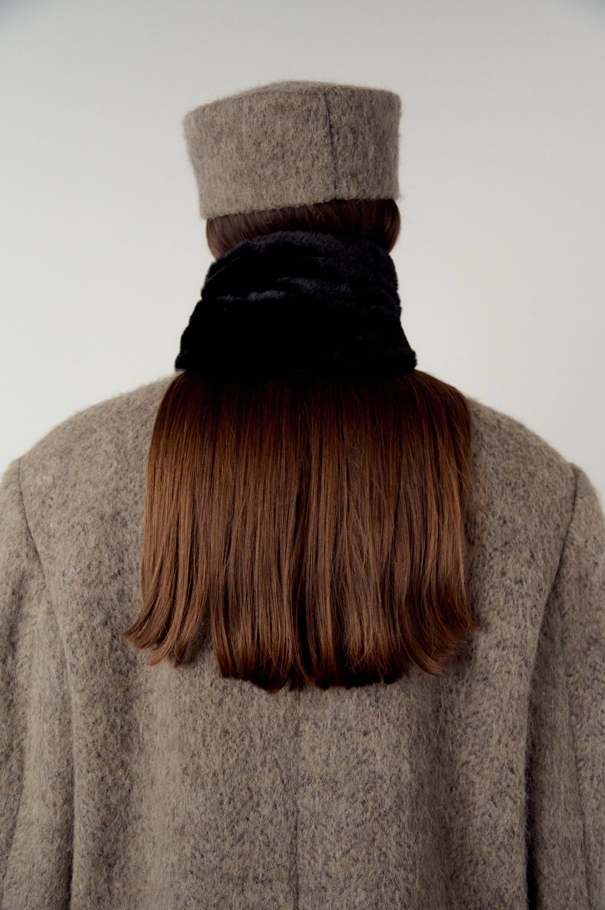Woman wearing a brown, warm grey coat and hat, and black scarf wrapped around hair on a plain white background