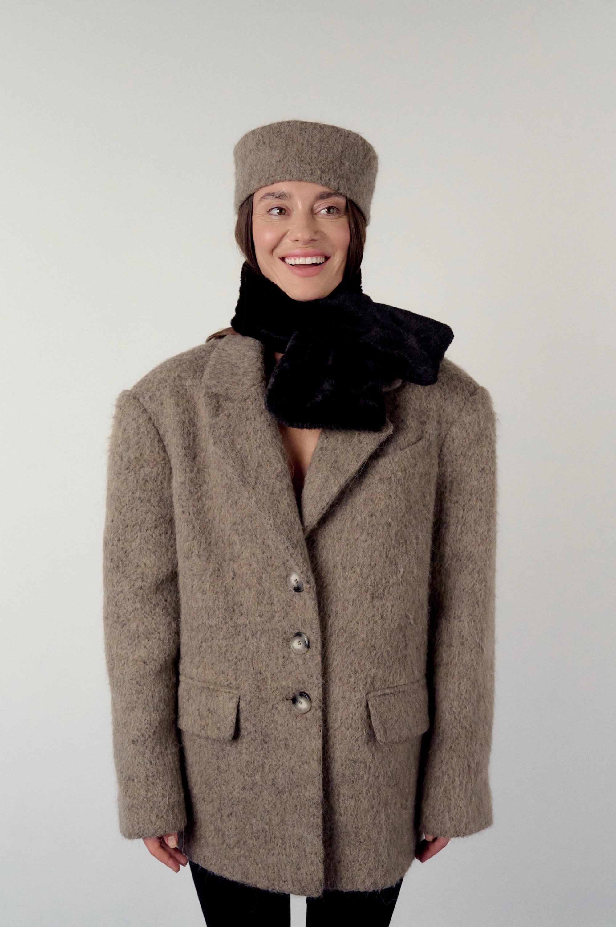 Woman wearing a brown, warm grey coat, black scarf, and hat on a white background